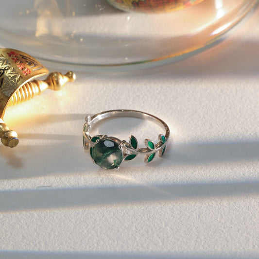 May include: A silver ring with a large, round, green gemstone at the center. The ring features a leaf design with green enamel accents. The ring is set on a white surface, with soft lighting.