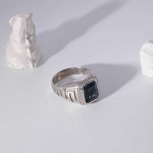 May include: A silver ring with a large, square, dark blue gemstone. The ring has a Greek key design on the sides and is set against a white background with decorative white stone sculptures.