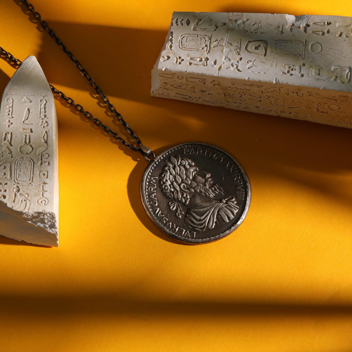 May include: A silver-colored pendant necklace featuring a detailed coin design with a profile of a man's face. The necklace is displayed on a yellow surface, alongside two white, carved stone fragments with hieroglyphic-like inscriptions.