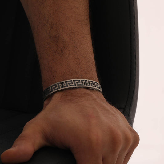 May include: A silver-toned cuff bracelet featuring a continuous black Greek key pattern engraved along the outer surface. The metal band has a textured finish and a wide, flat profile that wraps around the wrist with a clean, geometric design.